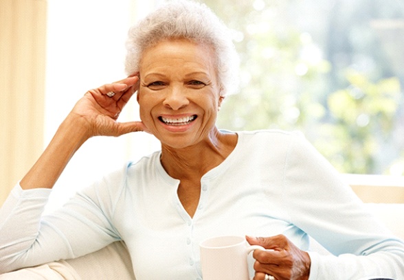 An older woman seated on a couch and holding a cup in her hand while smiling after seeing a dentist near Milford for comprehensive dental care