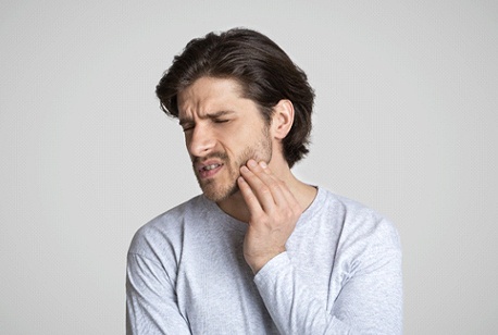 A young man wearing a gray shirt holds his cheek in pain, needing an emergency dentist